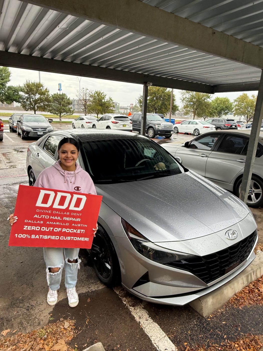 Customer holding a Divine Dallas Dent sign beside a silver Hyundai Elantra after hail repair